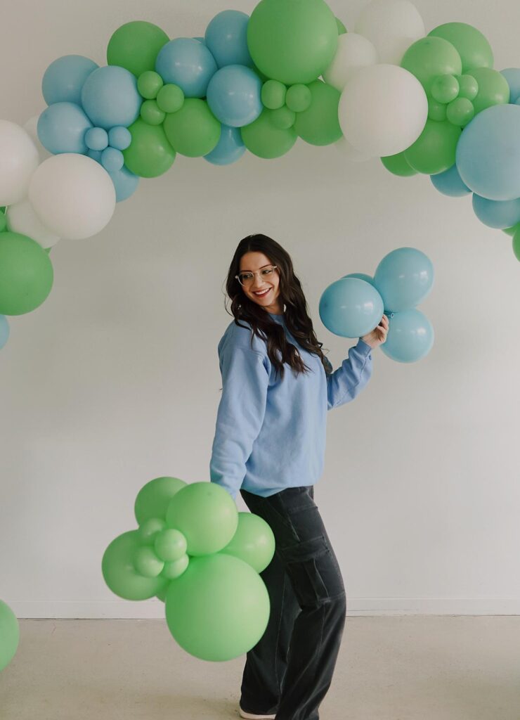 Alexis stands under a green, blue and white balloon arch while holding cute clumps of blue and green balloons in her hands.