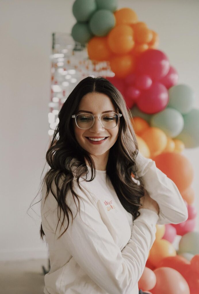 Alexis looks to the side with her hand in her hair while she stands in front of a green, orange, red, and pink balloons.