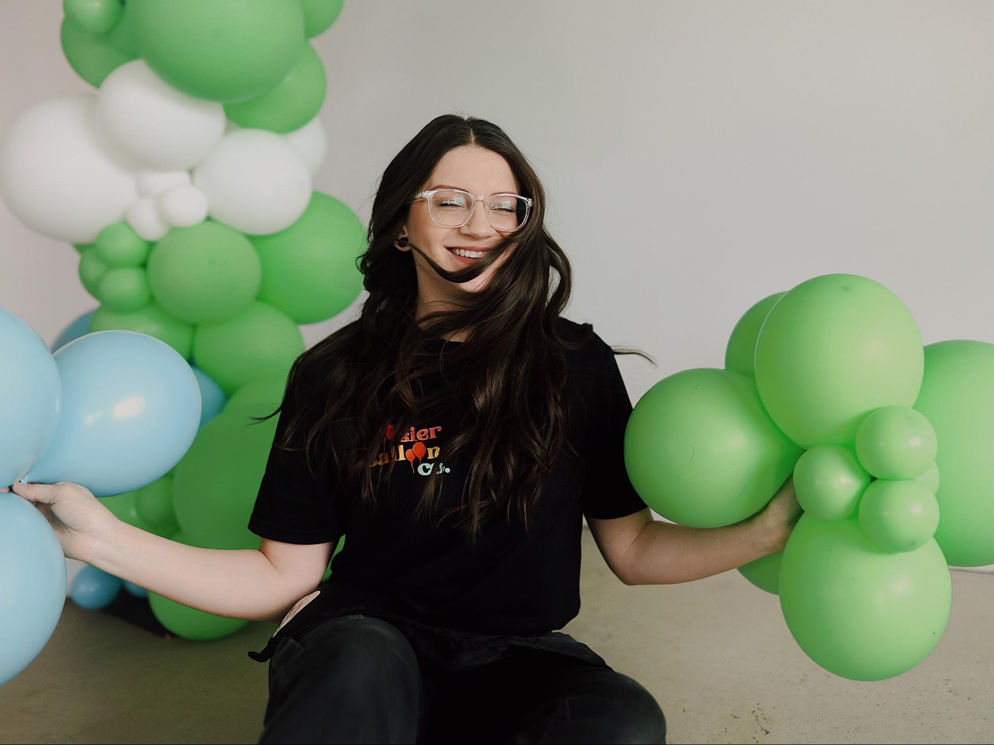Alexis sits holding cute clumps of balloons in her hands, green in her left and blue in her right. She is sitting in front of a green and white balloon arch.