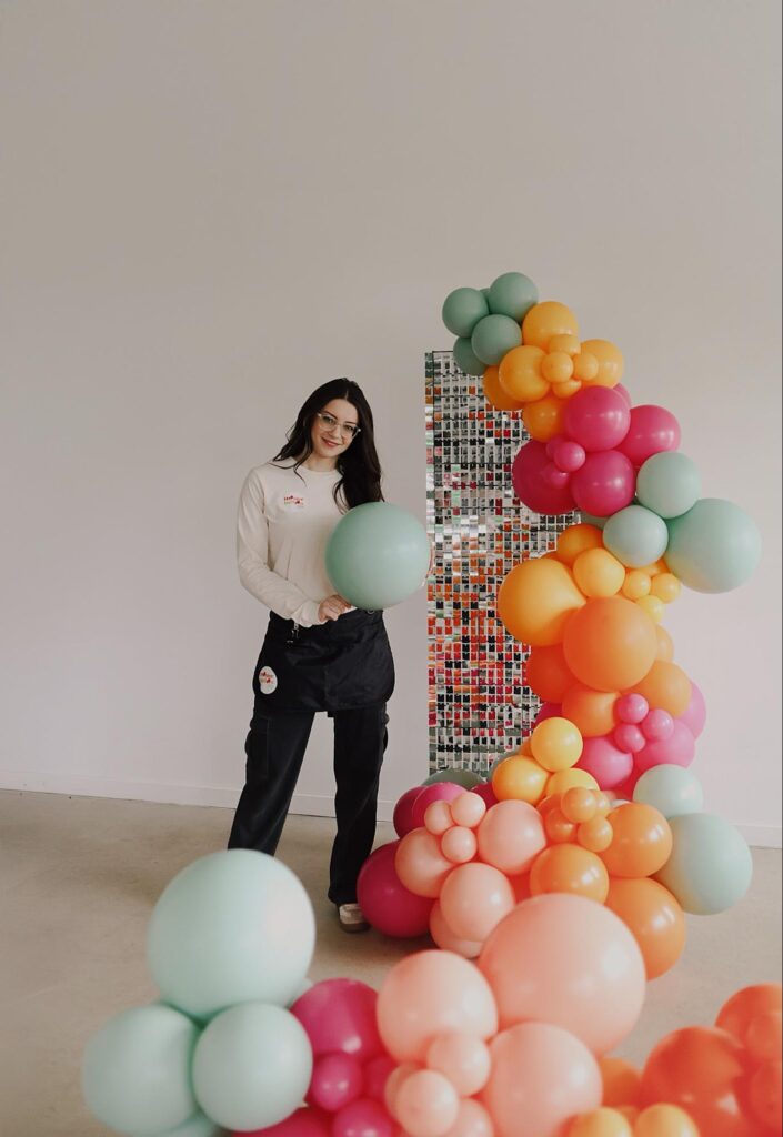 Alexis stands next to a cascade of green, orange, red, and pink balloons while she holds a green balloon.