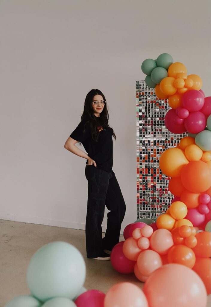 Alexis stands next to a cascade of green, orange, red, and pink balloons.