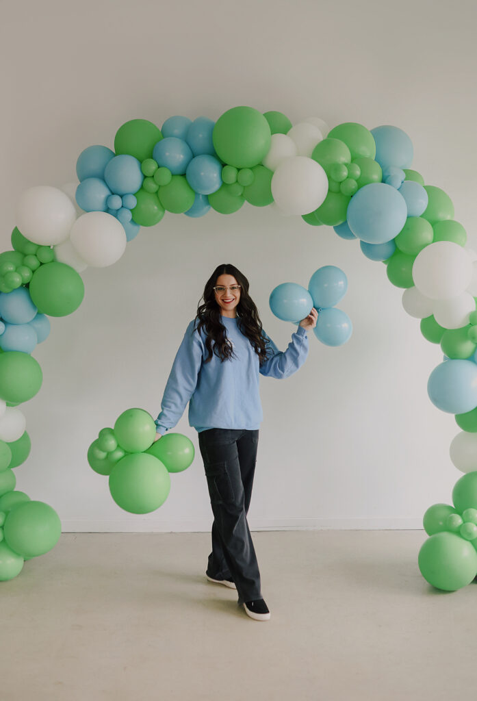 Alexis stands under a green, blue, and white balloon arch with cute clumps of green and blue balloons in her hands.
