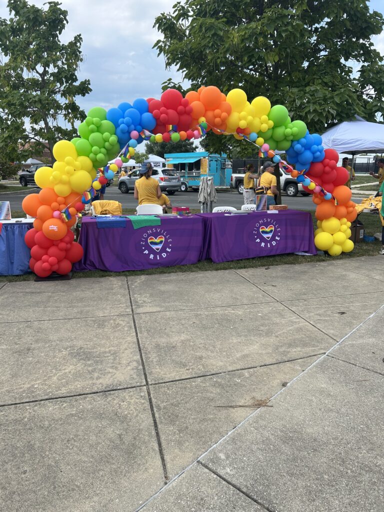 A rainbow balloon arch is in front of two long tables with purple tablecloths.