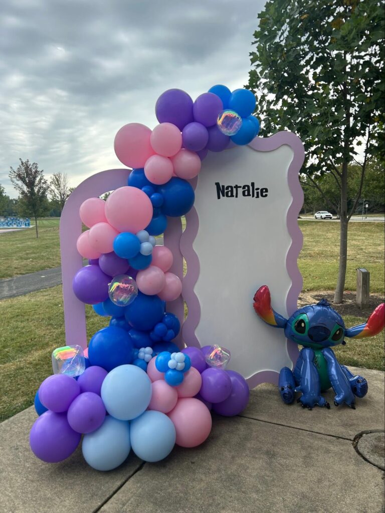 A cascade of purple, pink, light blue and dark blue balloons are on the left side of a rounded backdrop that says Natalie. There is a balloon image of Stitch on the right.