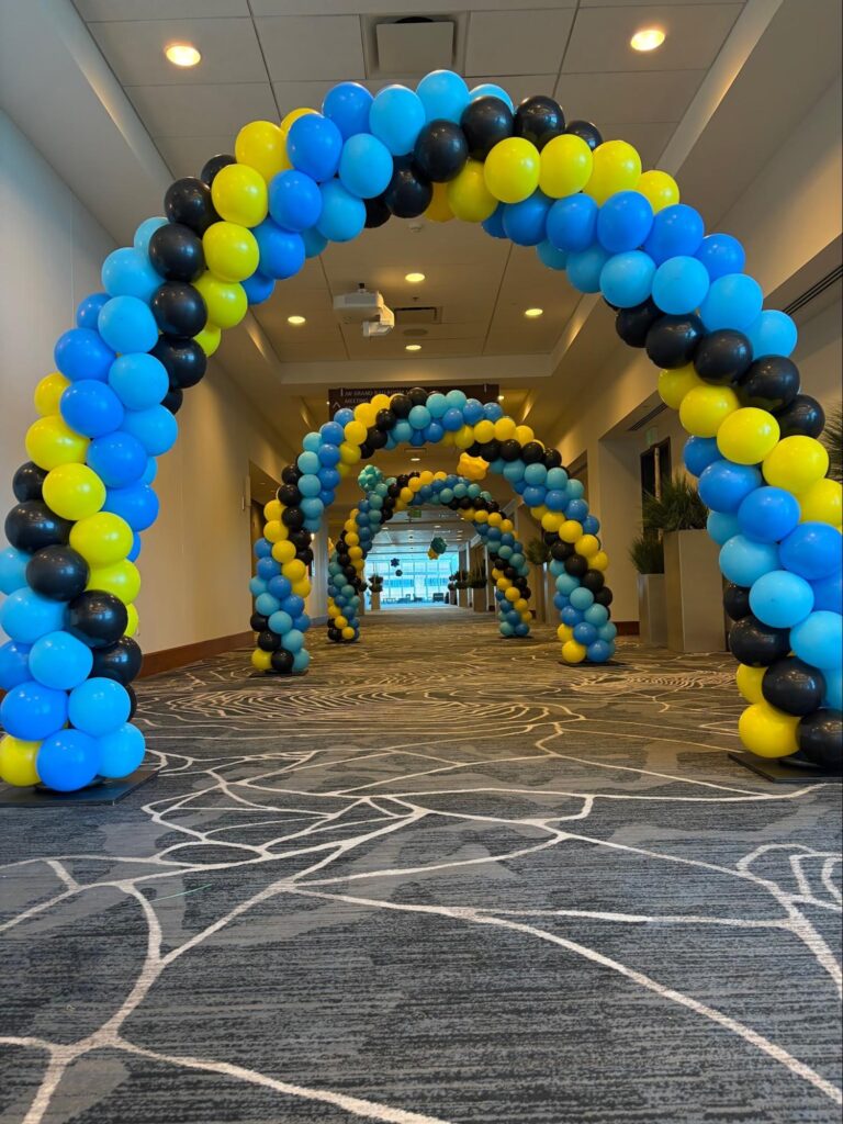 Multiple balloon arches of blue, yellow, and black line a long and large hallway.