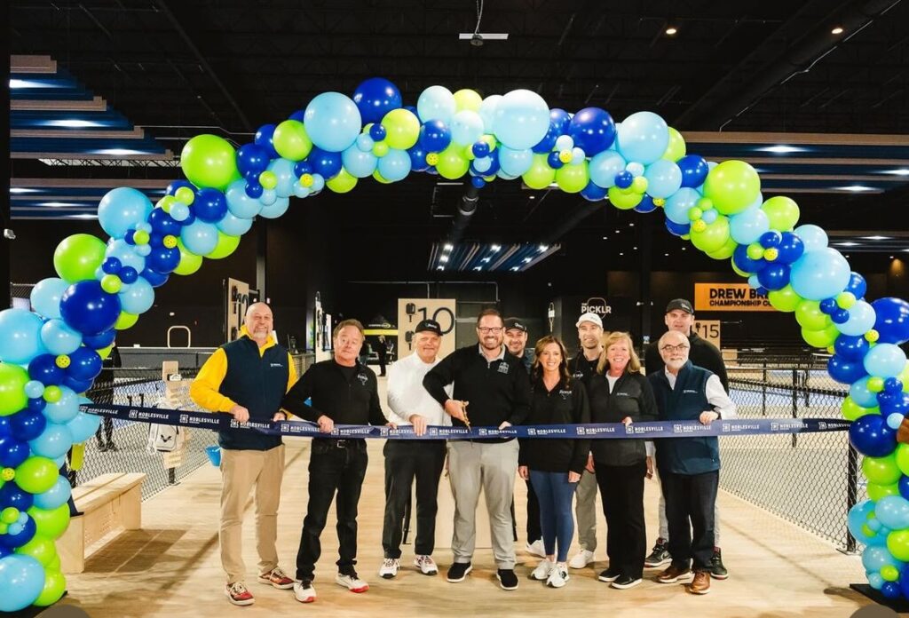 A group of people stand under a balloon arch ready to cut a ribbon for a grand opening.