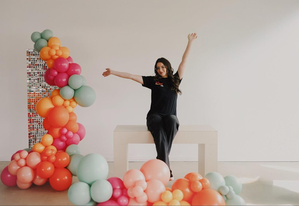 Alexis sits with her arms up surrounded by pink, red, orange, and light blue balloons.