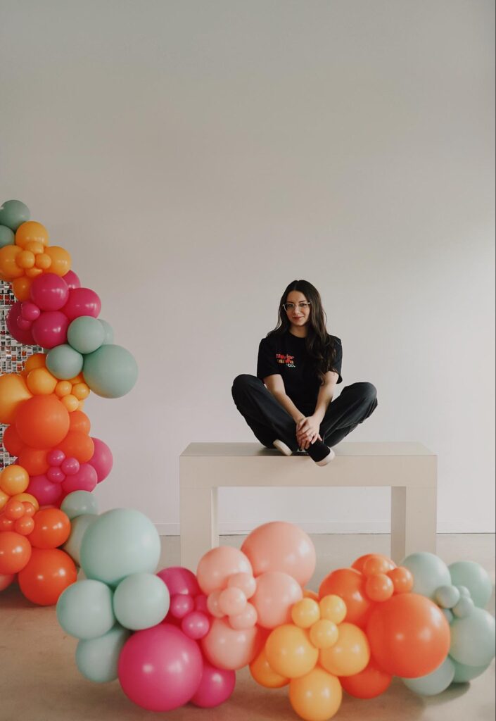 Alexis sits on a bench surrounded by a large balloon design of light green, orange, red, and pink balloons.