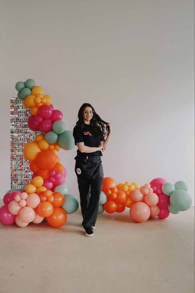 Alexis stands in front of a balloon display with orange, green, red and pink balloons. Her hair looks like it's blowing in the wind.