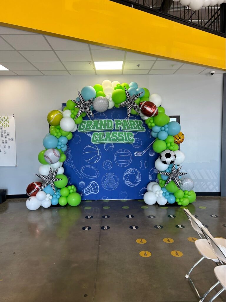 A balloon arch with white, light blue,  green, football, soccer, basketball and race balloons frames a blue photo op background with the words Grand Park Classic in green.