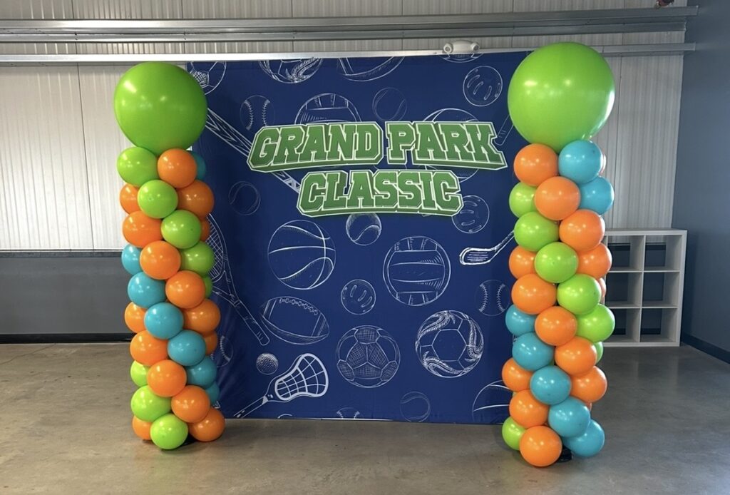 Two orange, green, and blue balloon columns with large green balloons on top stand on the edges of a blue photo op background with the words Grand Park Classic in green and sports balls and equipment graphics in white.
